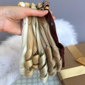 May include: A hand holding five swatches of doll hair in varying shades of blonde and brown. The swatches are labeled 1 through 5, with the first being a light blonde and the last a deep auburn. The hair is styled in loose curls.