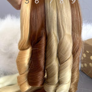 May include: A hand holds several swatches of hair extensions in various shades of blonde and brown. The hair samples are numbered 5 through 9. The colors range from light blonde to dark brown, with each swatch showing a curled texture.