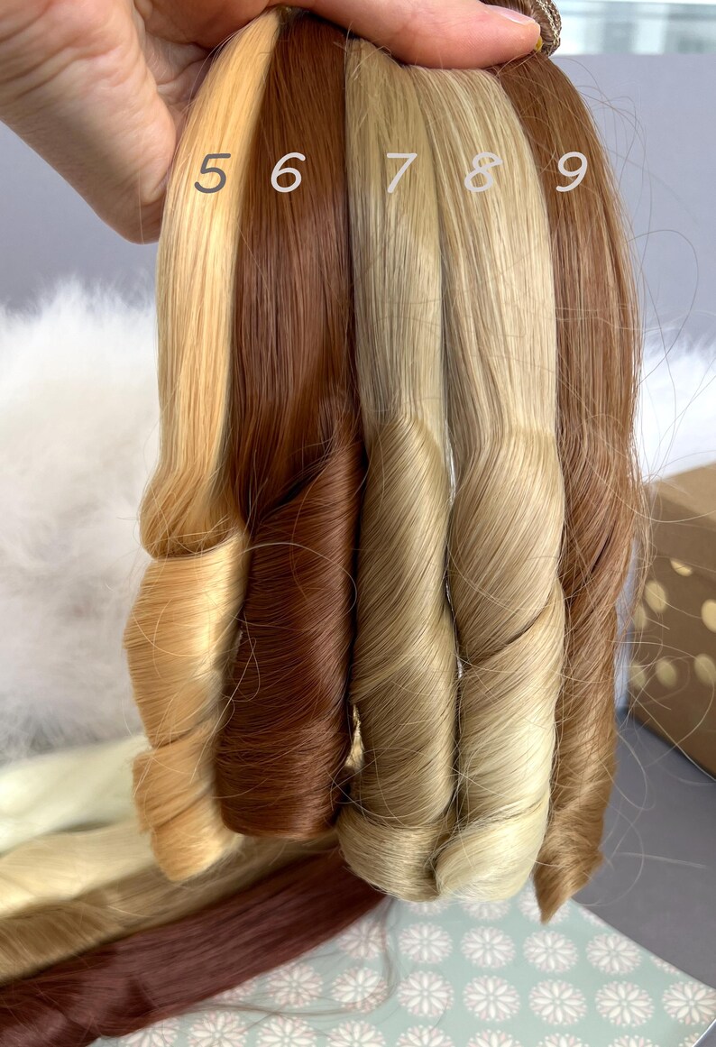 May include: A hand holds several swatches of hair extensions in various shades of blonde and brown. The hair samples are numbered 5 through 9. The colors range from light blonde to dark brown, with each swatch showing a curled texture.