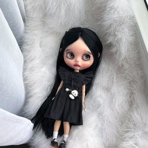 May include: A doll with long straight black hair, large brown eyes, and pale skin. The doll wears a black sleeveless dress with ruffled shoulders, a small white panda charm attached to the waist, white socks, and black shoes with white laces.
