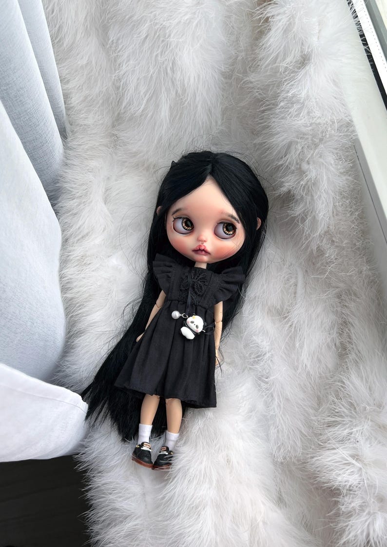 May include: A doll with long straight black hair, large brown eyes, and pale skin. The doll wears a black sleeveless dress with ruffled shoulders, a small white panda charm attached to the waist, white socks, and black shoes with white laces.