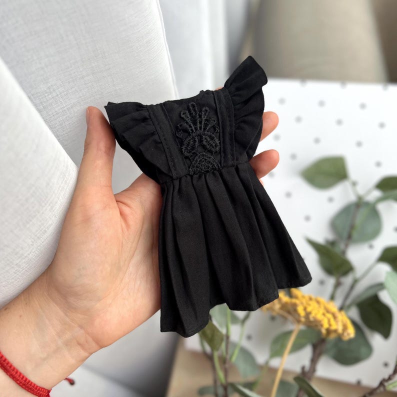 May include: A small black dress featuring ruffled sleeves and a central vertical panel with intricate black lace embroidery. The garment has a pleated skirt section and a structured bodice, designed with a simple round neckline and a solid black fabric.