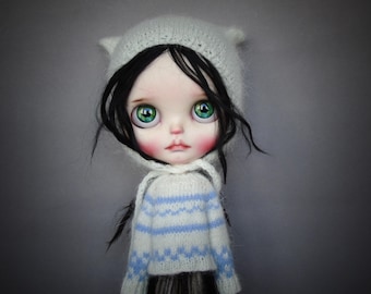 May include: A doll with large green eyes, pale skin, and dark hair, wearing a white knit hat with small ear-like protrusions and a white long-sleeved sweater featuring horizontal light blue stripes and a geometric pattern across the chest and sleeves.