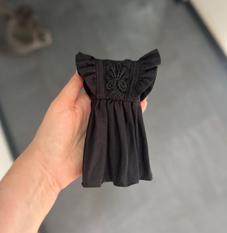 May include: A small black dress for a doll featuring ruffled cap sleeves and a central vertical panel decorated with intricate black lace embroidery. The garment has a gathered waistline and a simple skirt, showcasing detailed stitching throughout.
