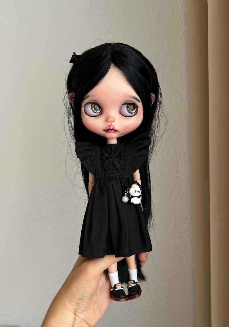 May include: A doll with long straight black hair, large brown eyes, and pale skin. The doll wears a black dress with ruffled sleeves and a small panda-shaped bag with a pearl charm. It also wears white socks and black shoes with brown soles.