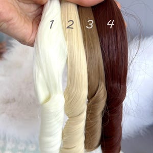 May include: Four bundles of hair extensions in varying shades of blonde and brown. The colors are numbered 1 through 4, with the first being a light blonde, followed by a medium blonde, a light brown, and a dark auburn. The extensions are held together at the top.