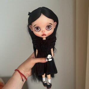 May include: A doll with long straight black hair, large brown eyes, and pale skin. The doll wears a black dress with ruffled shoulders and a small panda charm accessory. It also wears white socks and black shoes with white details on the front.