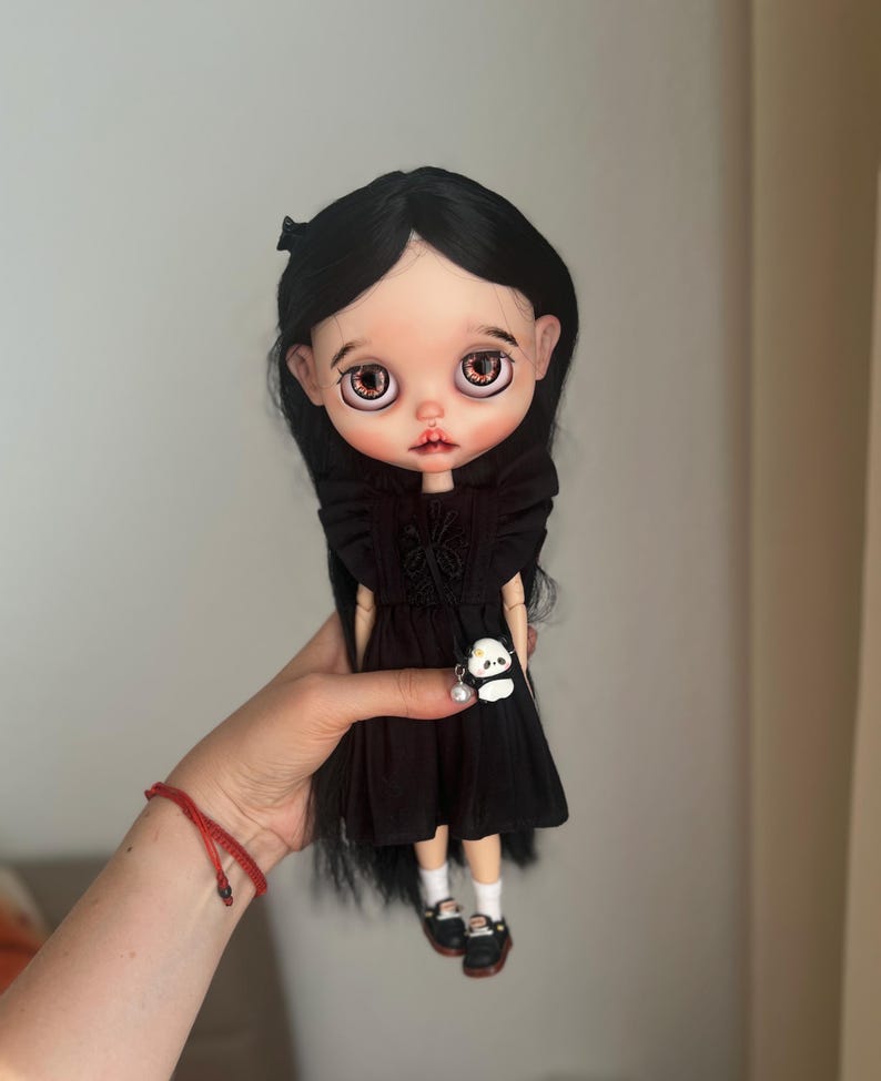 May include: A doll with long straight black hair, large brown eyes, and pale skin. The doll wears a black dress with ruffled shoulders and a small panda charm accessory. It also wears white socks and black shoes with white details on the front.