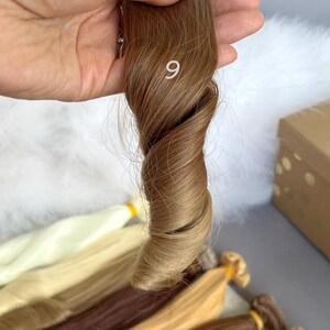 May include: A sample of light brown hair extensions, held in a hand, with the number 9 visible. Several other bundles of hair extensions in various shades of blonde and brown are visible in the background.