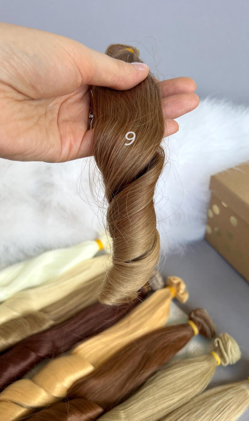 May include: A sample of light brown hair extensions, held in a hand, with the number 9 visible. Several other bundles of hair extensions in various shades of blonde and brown are visible in the background.