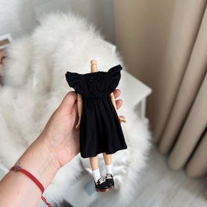 May include: A doll wearing a black dress with ruffled sleeves and intricate embroidery on the bodice. The doll has white socks and black shoes with white accents. The dress features a gathered skirt and a round neckline, providing a detailed look at the miniature clothing and footwear.