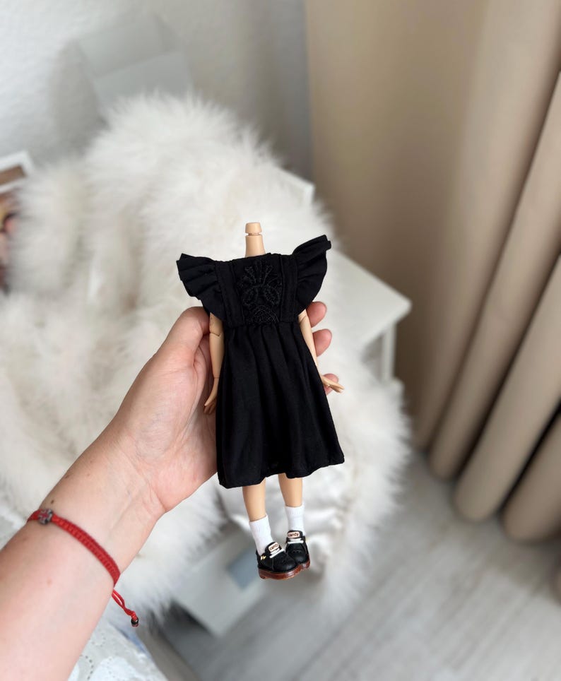 May include: A doll wearing a black dress with ruffled sleeves and intricate embroidery on the bodice. The doll has white socks and black shoes with white accents. The dress features a gathered skirt and a round neckline, providing a detailed look at the miniature clothing and footwear.