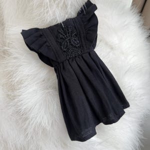 May include: A black dress featuring ruffled sleeves and a central vertical panel with intricate black floral embroidery. The garment has a gathered waistline that flows into a pleated skirt, creating a structured yet fluid silhouette.