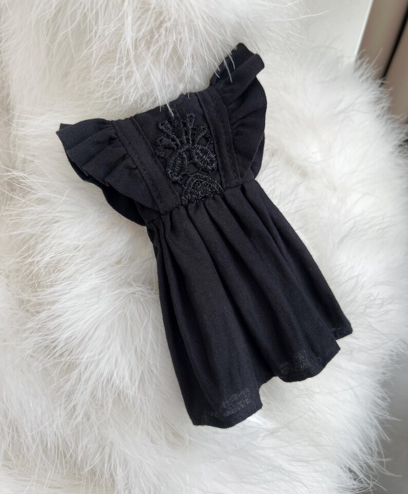 May include: A black dress featuring ruffled sleeves and a central vertical panel with intricate black floral embroidery. The garment has a gathered waistline that flows into a pleated skirt, creating a structured yet fluid silhouette.