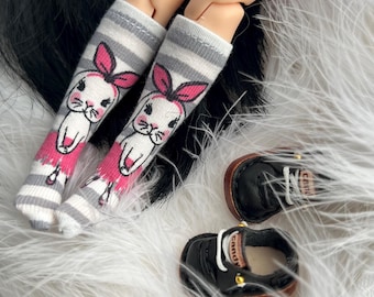 May include: Doll socks with a gray and white striped pattern, featuring a cartoon bunny design with pink accents. Two small black doll shoes with white laces and brown soles are also visible.