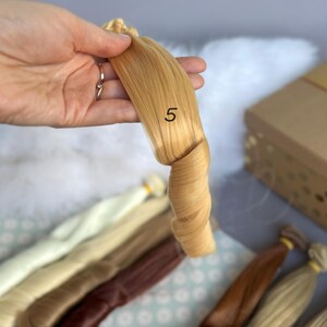 May include: A bundle of light blonde doll hair, labeled with the number "5", is held up against a backdrop of other hair colors. The hair is tightly curled at the end. Other hair bundles in various shades of blonde and brown are visible.