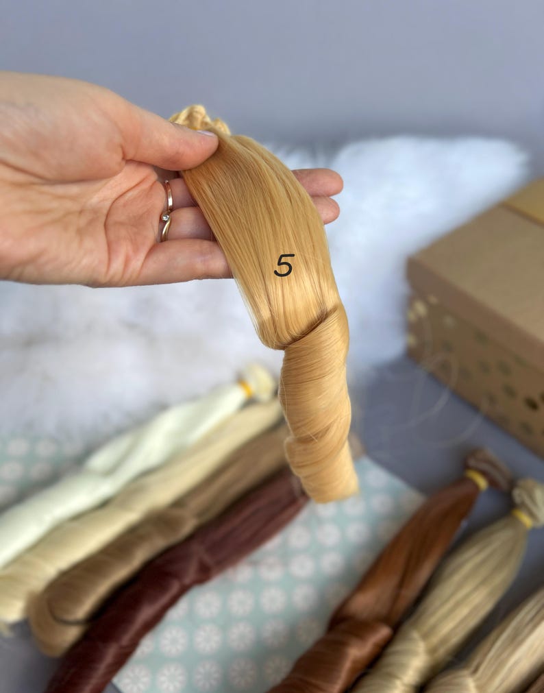 May include: A bundle of light blonde doll hair, labeled with the number "5", is held up against a backdrop of other hair colors. The hair is tightly curled at the end. Other hair bundles in various shades of blonde and brown are visible.