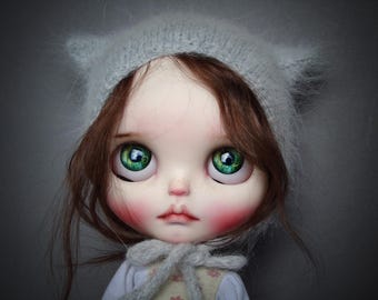 May include: A doll with large, expressive green eyes and rosy cheeks. The doll has brown hair and wears a light gray hat with pointed ears. A gray scarf is tied around the neck. The doll's face has a delicate, painted appearance.