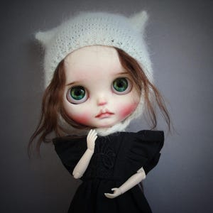 May include: A doll with large green eyes, rosy cheeks, and brown hair wearing a white knitted hat with small pointed ears. The doll is dressed in a black garment with ruffled sleeves and a textured bodice, featuring articulated arms and hands.