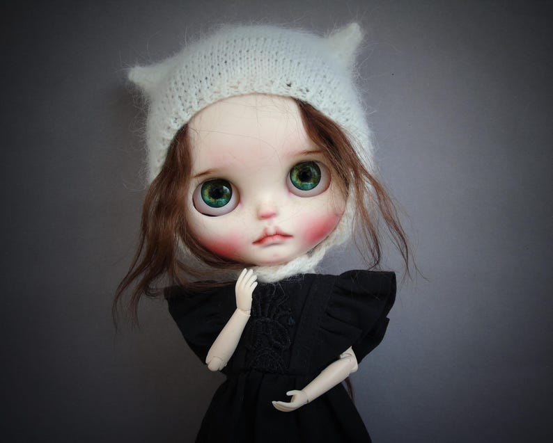 May include: A doll with large green eyes, rosy cheeks, and brown hair wearing a white knitted hat with small pointed ears. The doll is dressed in a black garment with ruffled sleeves and a textured bodice, featuring articulated arms and hands.