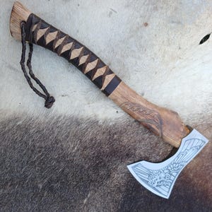 May include: A handcrafted axe with a wooden handle and a decorative blade. The handle features a wrapped grip with a leather cord. The blade has an etched design. The axe is displayed on a textured surface.
