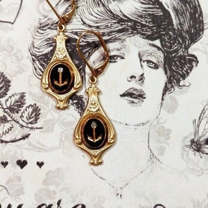 May include: Gold tone earrings with black enamel and a small anchor charm. The earrings have a teardrop shape and are accented with a clear rhinestone.