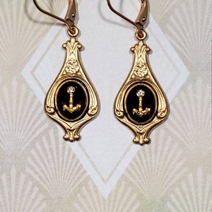 May include: Gold dangle earrings with black enamel and a gold anchor charm. The earrings have a floral design and a small clear rhinestone on the anchor.
