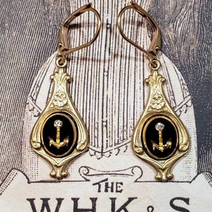 May include: Gold dangle earrings with black enamel and a gold anchor charm. The anchor is set with a small clear rhinestone.