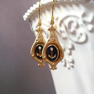 May include: Gold dangle earrings with a black enamel anchor charm and a single rhinestone. The anchor is set in a gold oval frame.