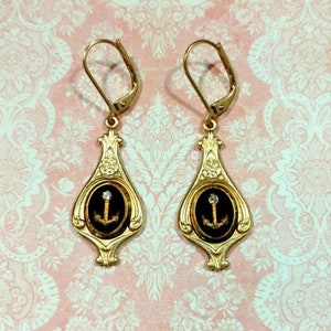 May include: Gold dangle earrings with black enamel and a gold anchor with a single rhinestone in the center. The earrings have a teardrop shape with a decorative design.