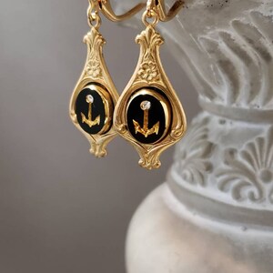 May include: Gold dangle earrings with black enamel and a gold anchor charm. Each earring has a single clear rhinestone.
