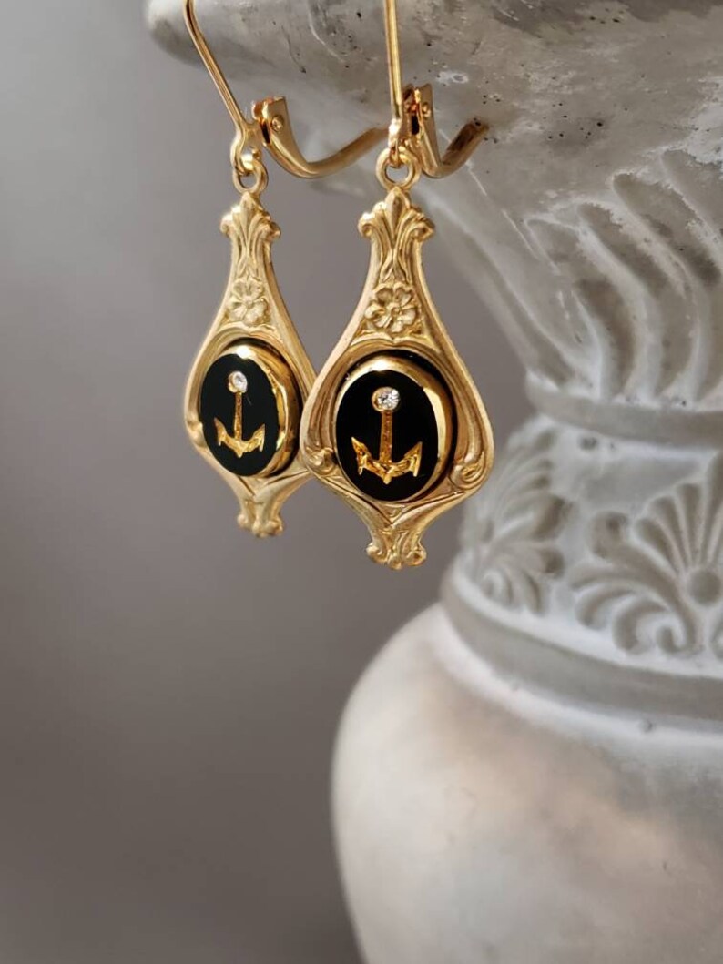 May include: Gold dangle earrings with black enamel and a gold anchor charm. Each earring has a single clear rhinestone.