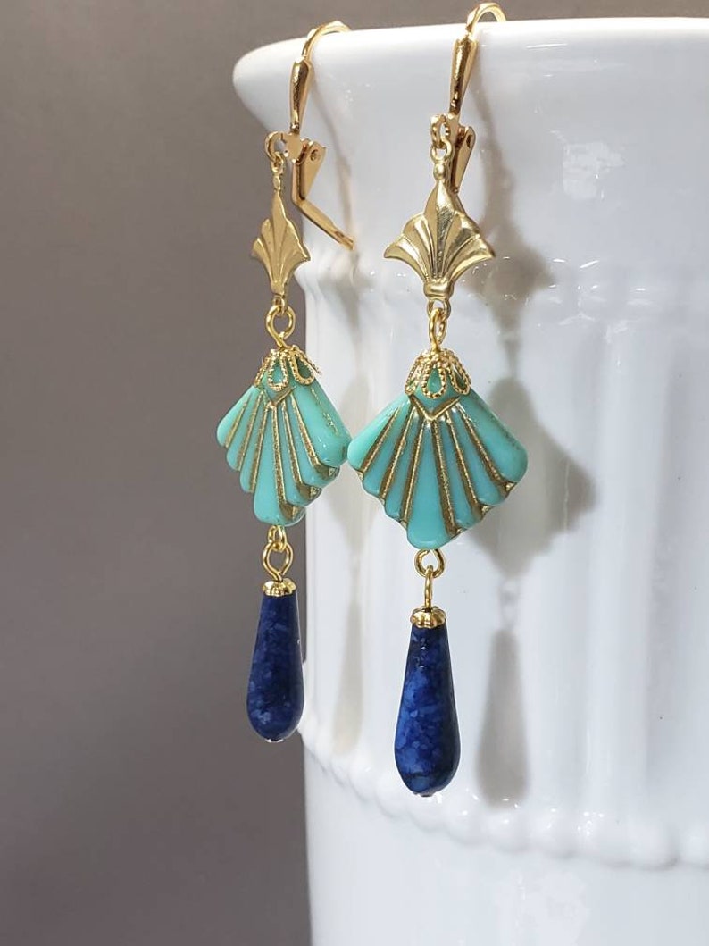May include: A pair of gold-tone drop earrings featuring a fan-shaped turquoise bead with gold detailing, topped with a gold metal fan accent, and finished with a dangling dark blue teardrop-shaped stone bead suspended from a gold-tone leverback.