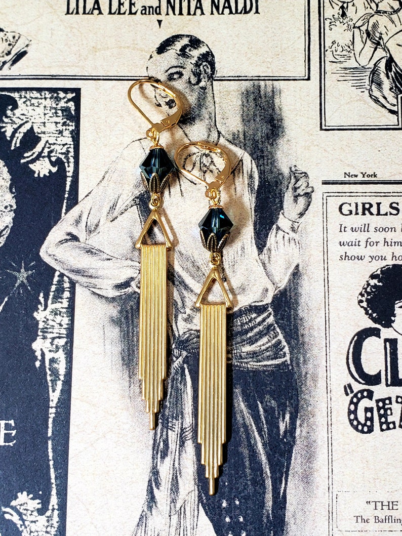K&ouml;nnte beinhalten: Ein Paar goldene Ohrringe mit einem dunkelblauen Kristallstein und einer Fransen aus goldenen St&auml;ben. Die Ohrringe h&auml;ngen vor einem Vintage-Hintergrund.