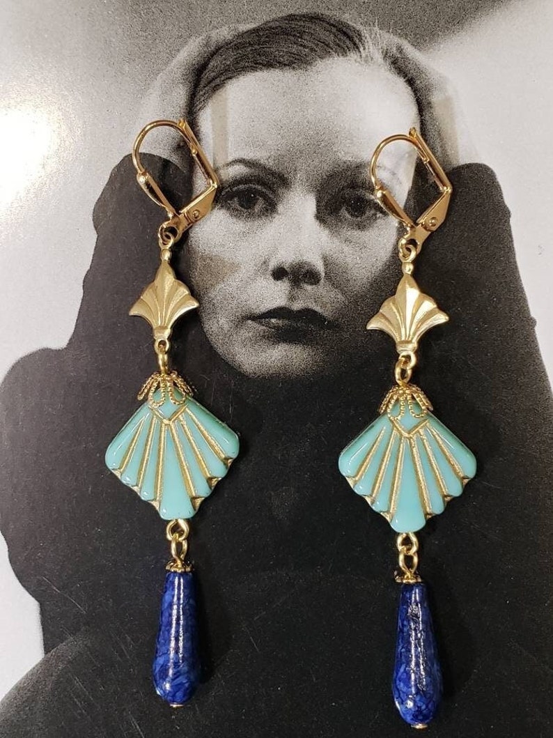 May include: A pair of gold-tone drop earrings featuring a fan-shaped turquoise enamel component with gold detailing, topped with a gold metal accent piece, and finished with a dangling, teardrop-shaped dark blue stone bead at the bottom.