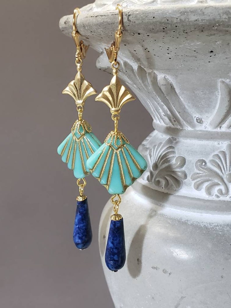 May include: A pair of gold-tone drop earrings featuring a fan-shaped turquoise-coloured component with gold-tone detailing, suspended from a gold-tone metal top piece, and finished with a dangling dark blue teardrop-shaped stone at the bottom.