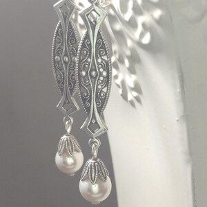 May include: A pair of silver-toned earrings with an ornate filigree design. Each earring features an elongated oval shape with intricate detailing and a dangling pearlescent bead. The earrings are suspended from a simple hook.