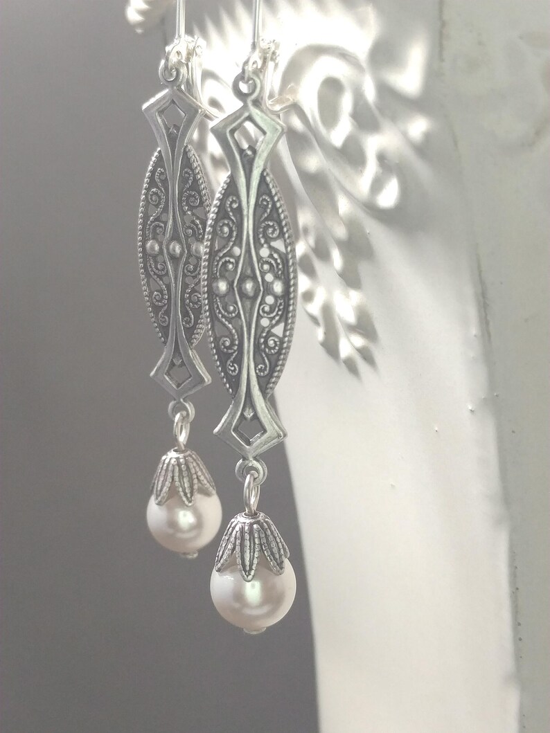 May include: A pair of silver-toned earrings with an ornate filigree design. Each earring features an elongated oval shape with intricate detailing and a dangling pearlescent bead. The earrings are suspended from a simple hook.