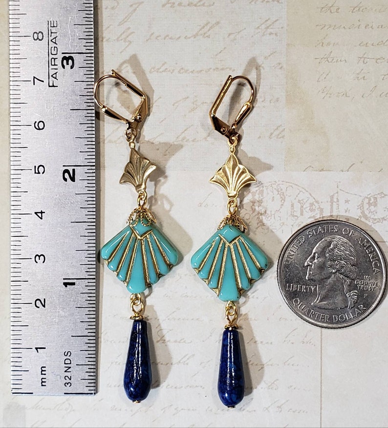 May include: A pair of gold-tone dangle earrings featuring a turquoise fan-shaped bead with gold accents and a dark blue teardrop-shaped stone at the base. A metal ruler shows the earrings are approximately 8.3 cm long next to a US quarter coin.
