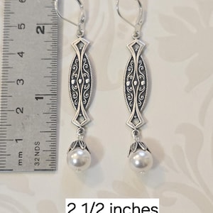 May include: A pair of silver-toned earrings with a decorative oval design and a white pearl drop. The earrings are 2 1/2 inches long. The design includes intricate scrollwork and a lever-back closure.