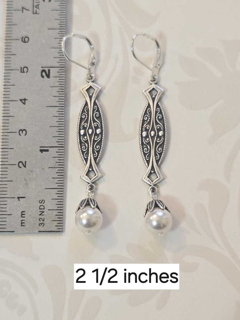 May include: A pair of silver-toned earrings with a decorative oval design and a white pearl drop. The earrings are 2 1/2 inches long. The design includes intricate scrollwork and a lever-back closure.
