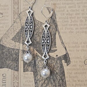 May include: Silver-toned dangle earrings with an ornate, elongated design and a pearlescent white pearl drop. The earrings feature a decorative filigree pattern and lever-back closures, perfect for adding a touch of vintage elegance.