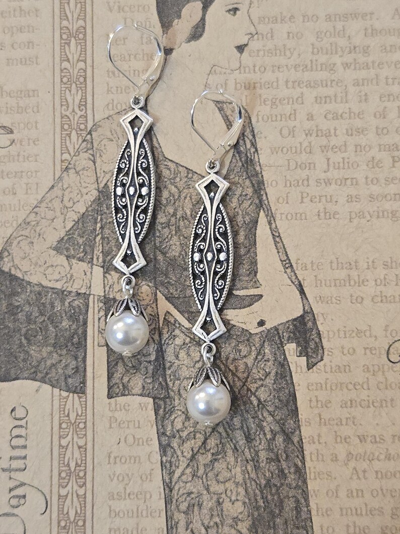 May include: Silver-toned dangle earrings with an ornate, elongated design and a pearlescent white pearl drop. The earrings feature a decorative filigree pattern and lever-back closures, perfect for adding a touch of vintage elegance.