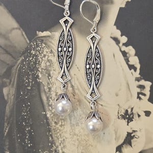 May include: A pair of silver-toned drop earrings. Each earring features an elongated, oval-shaped design with intricate scrollwork and a diamond-shaped accent. A pearlescent, round bead hangs from a decorative cap at the bottom. The earrings have lever-back closures.