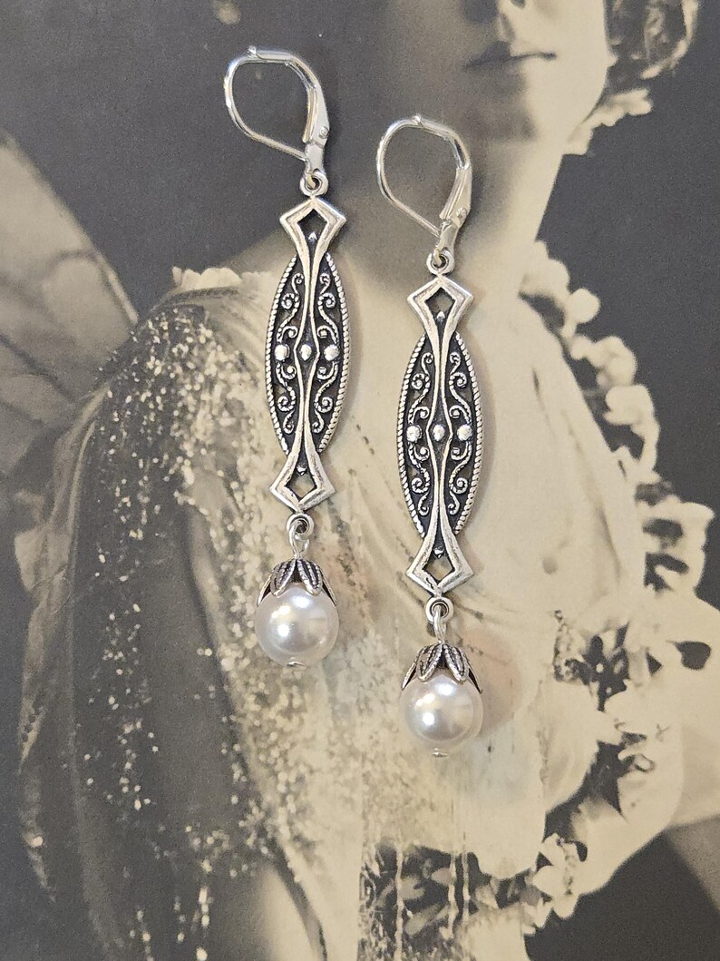 May include: A pair of silver-toned drop earrings. Each earring features an elongated, oval-shaped design with intricate scrollwork and a diamond-shaped accent. A pearlescent, round bead hangs from a decorative cap at the bottom. The earrings have lever-back closures.