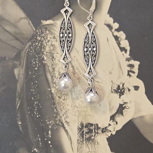 May include: Silver-toned dangle earrings with an ornate, elongated design. Each earring features a central oval with intricate scrollwork and a pearlescent bead drop. The earrings have lever-back closures.