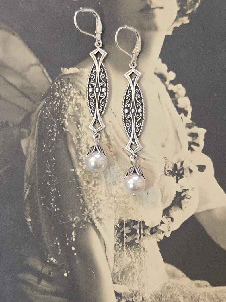 May include: Silver-toned dangle earrings with an ornate, elongated design. Each earring features a central oval with intricate scrollwork and a pearlescent bead drop. The earrings have lever-back closures.