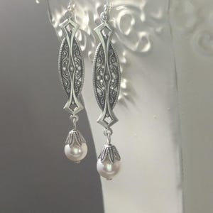 May include: Silver-toned dangle earrings with an ornate, elongated design. Each earring features a detailed filigree pattern and a teardrop-shaped, creamy white pearl. The earrings hang from delicate silver hooks.