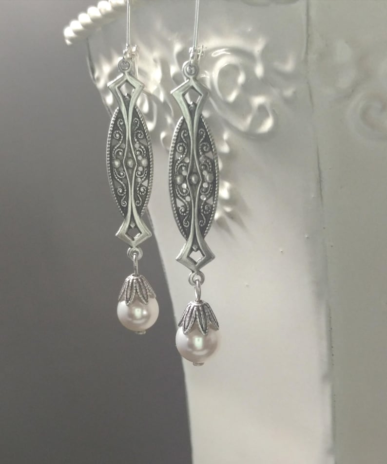 May include: Silver-toned dangle earrings with an ornate, elongated design. Each earring features a detailed filigree pattern and a teardrop-shaped, creamy white pearl. The earrings hang from delicate silver hooks.