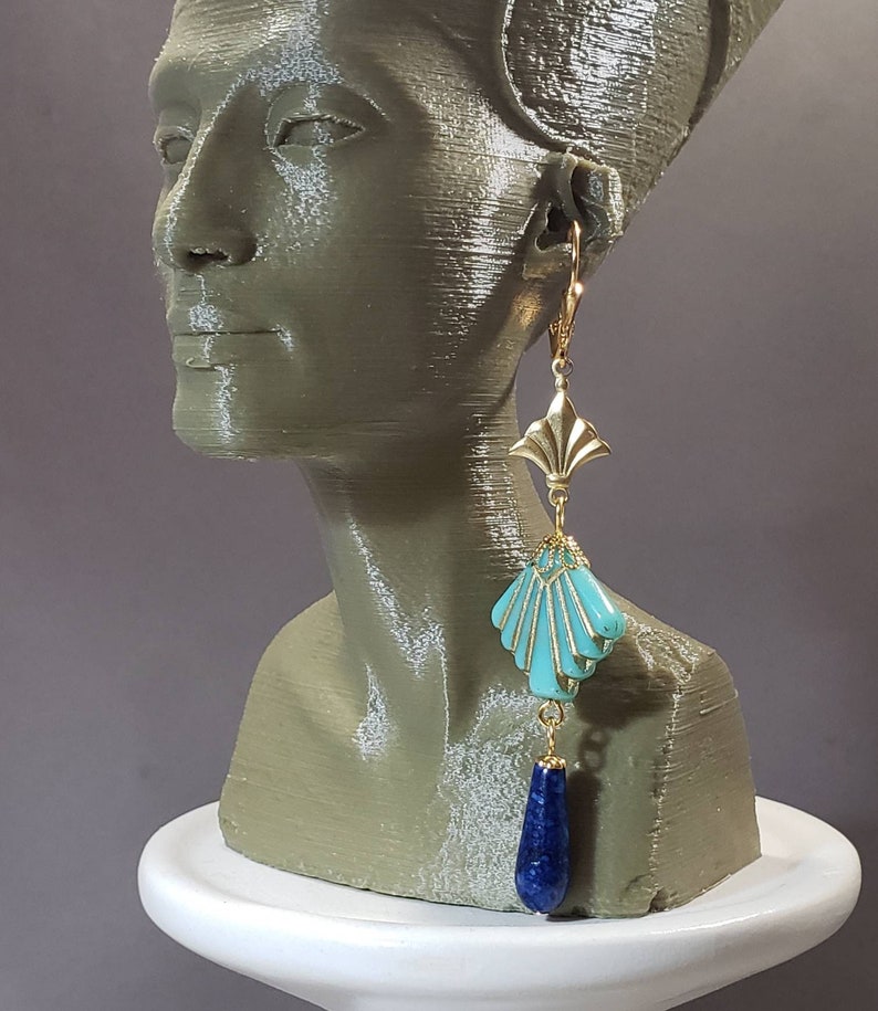 May include: A gold-toned leverback earring featuring a fan-shaped metal top, a central turquoise-coloured shell-shaped component with gold-toned filigree accents, and a dangling teardrop-shaped dark blue stone at the bottom of the earring.